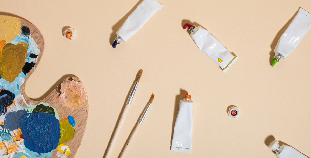 Photo facing down to a table top. On the table is a pallet covered in dry paint, paint brushes and tubes of coloured paint open with there caps spread around.