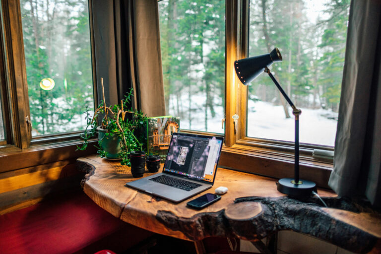 A desk in a home office. There is a plant, a laptop and a lamp on a wooden desk. The desk is by a window that shows snow and trees outside. The colours are warm.